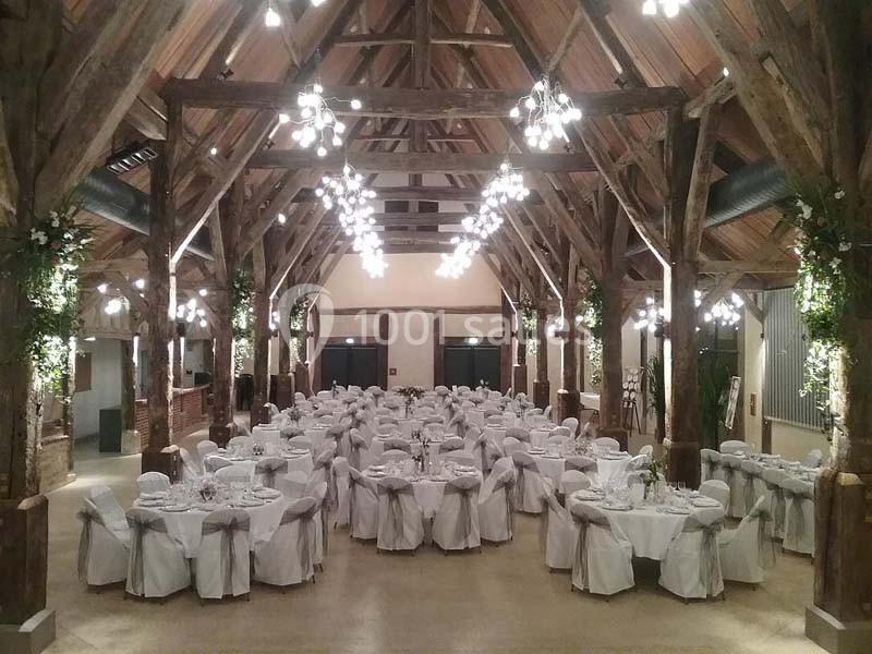 Salle de réception avec charpente en bois, tables rondes dressées et décorations lumineuses suspendues.