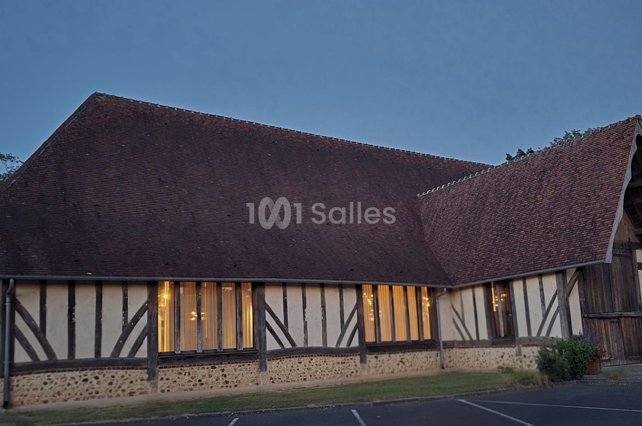 Bâtiment à colombages avec toit en pente, éclairé de l'intérieur, entouré de végétation et d'un parking.