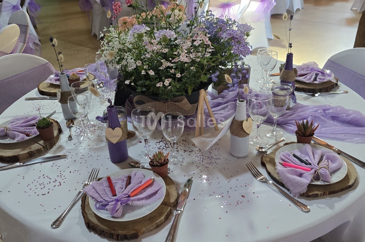Table décorée pour un événement avec nappes violettes, fleurs, bougies, rondins de bois et couverts disposés.