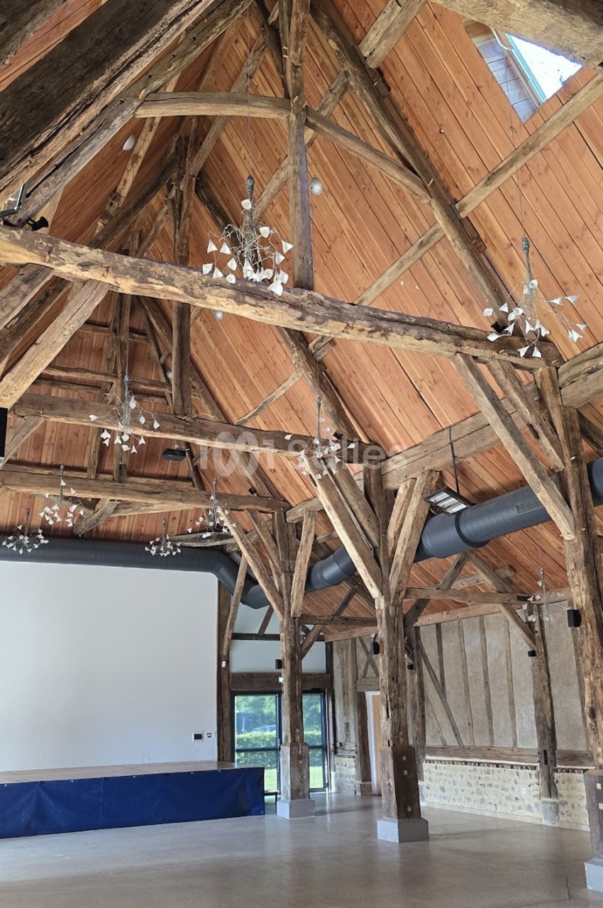 Charpente en bois apparente dans une grande salle lumineuse avec murs en pierre et écran blanc au fond.