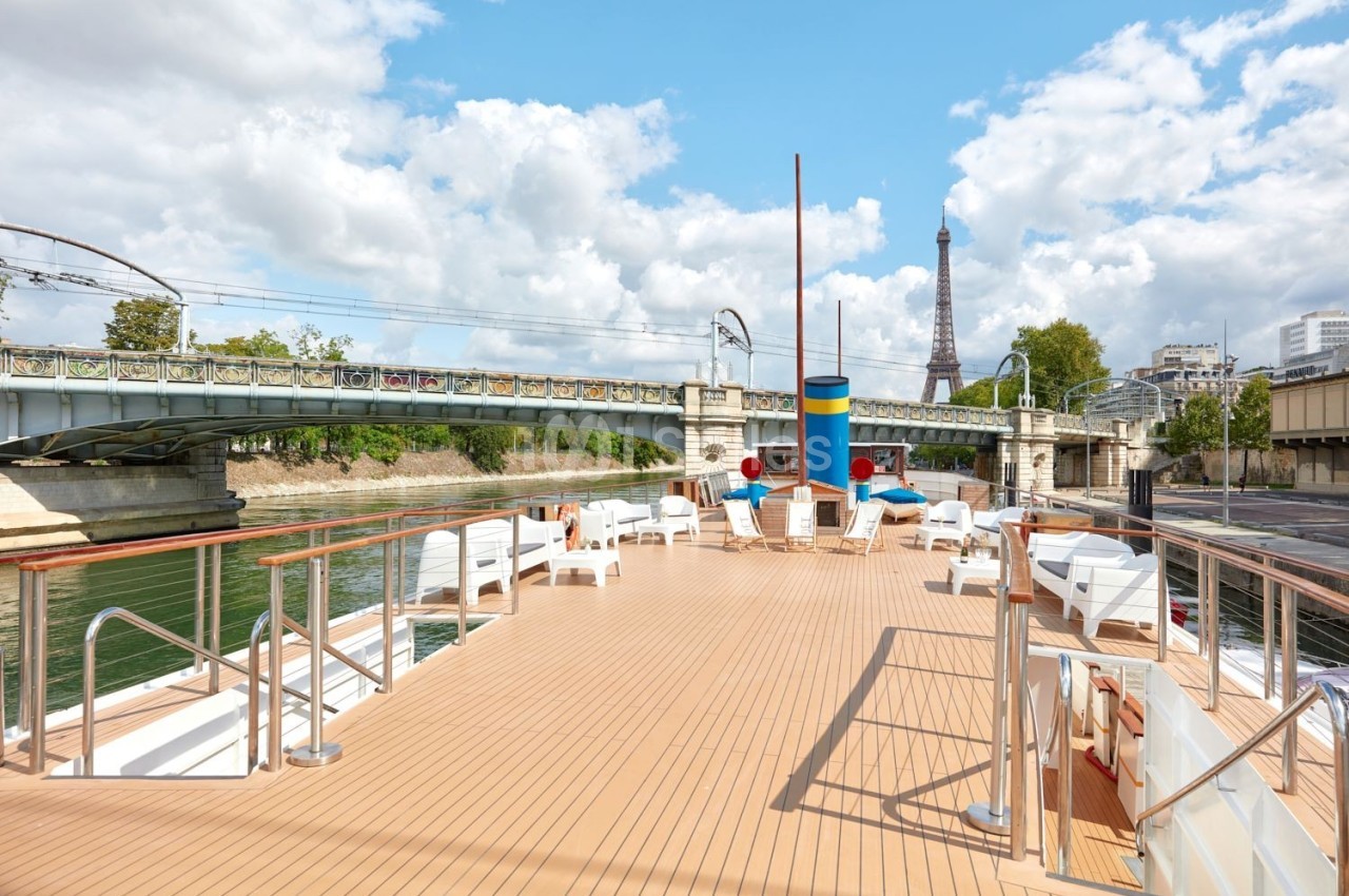 Pont supérieur d'un bateau sur la Seine avec des chaises longues, vue sur un pont et la tour Eiffel en arrière-plan.