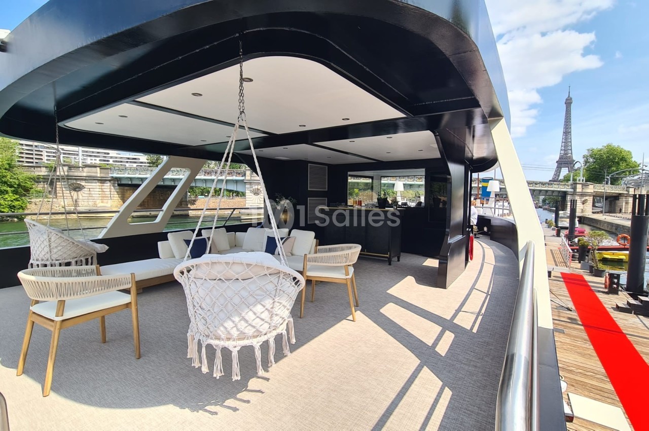 Terrasse d'un bateau avec salon extérieur, fauteuil suspendu, vue sur la Seine et la tour Eiffel en arrière-plan.