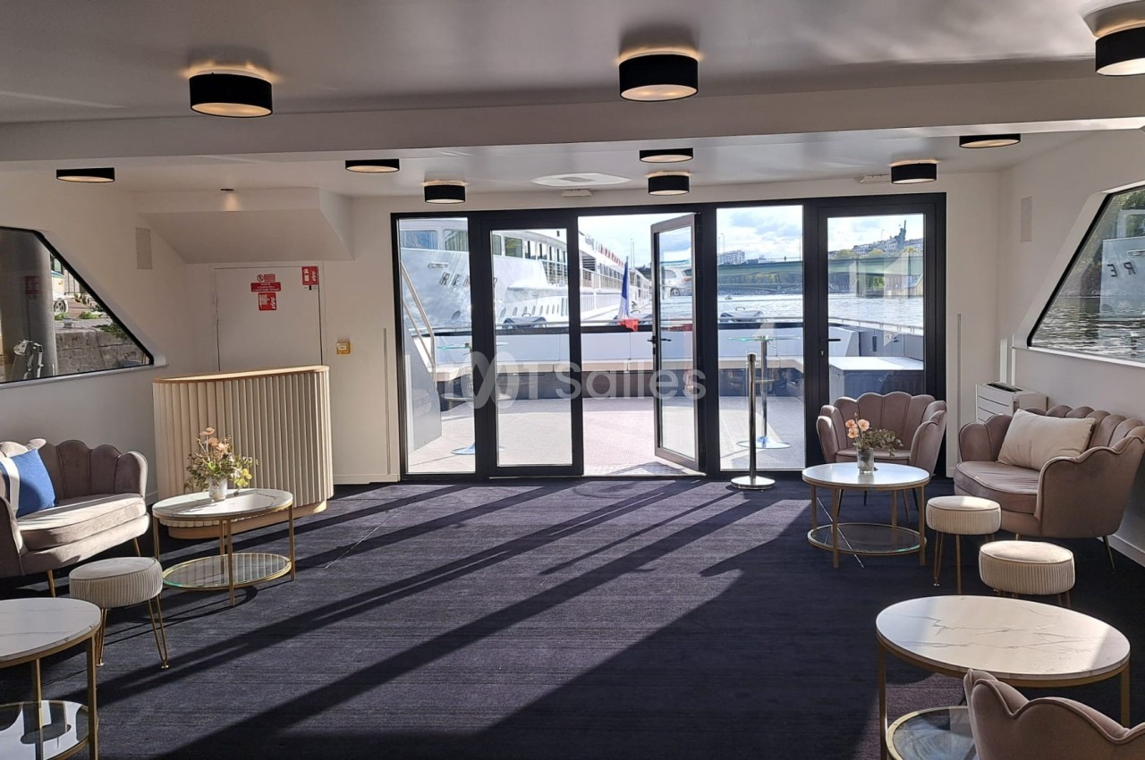 Salon lumineux avec fauteuils, tables basses et grandes baies vitrées donnant sur un quai et un bateau amarré.