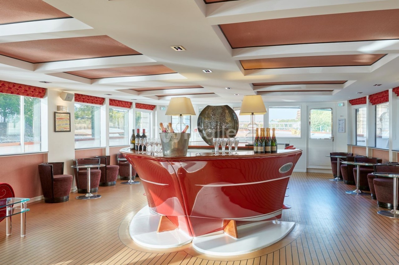 Bar moderne avec comptoir rouge brillant, bouteilles de champagne et verres, situé dans une salle lumineuse avec grandes…