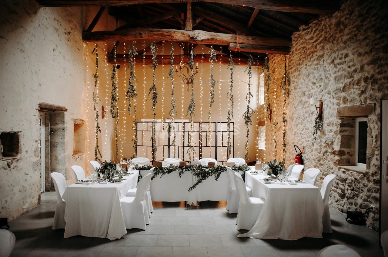 Salle de réception décorée avec des guirlandes lumineuses, des tables blanches et des arrangements floraux.