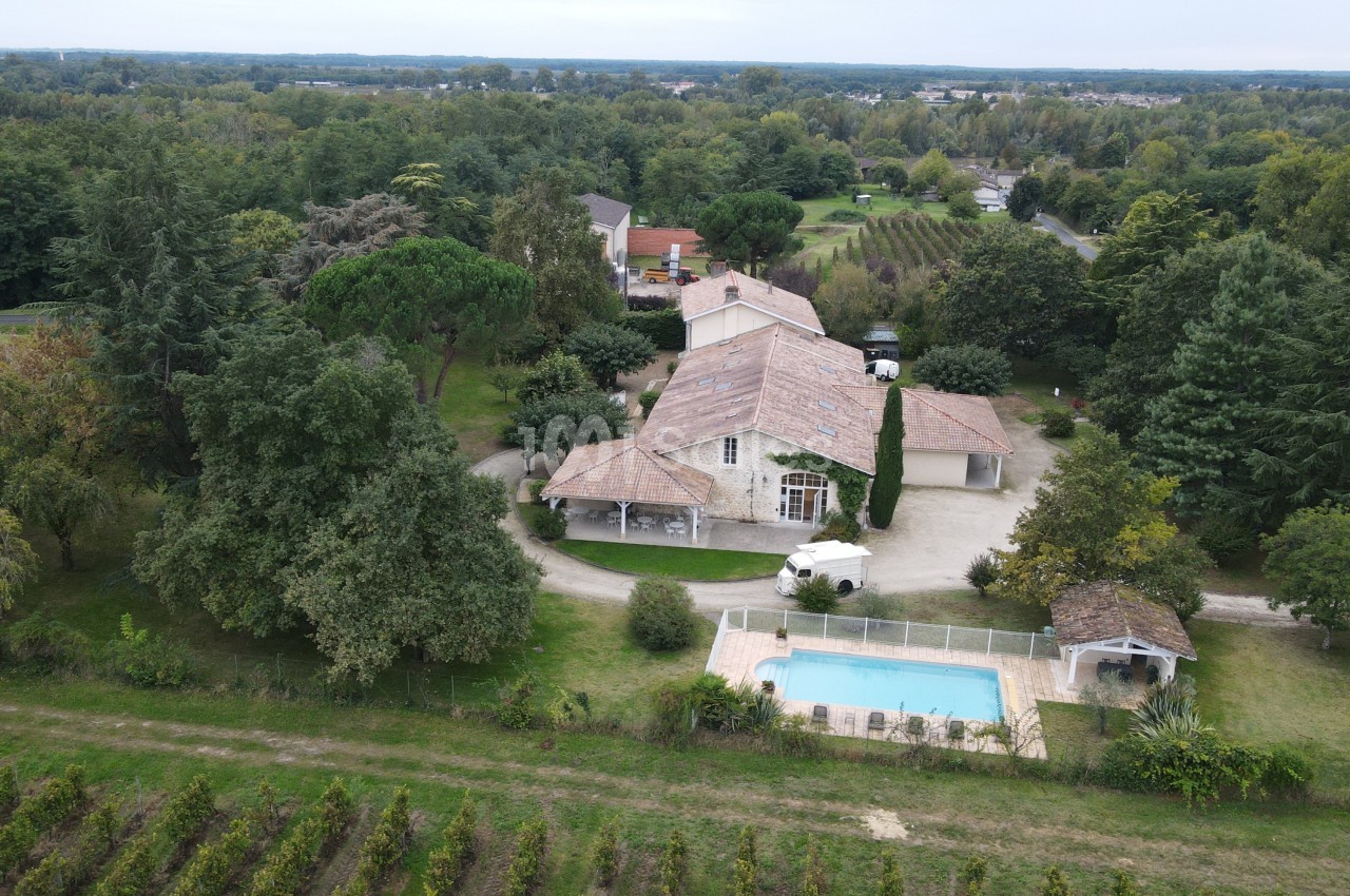 Vue aérienne d'une maison avec toit en tuiles, piscine clôturée, jardin arboré et vignes en arrière-plan.
