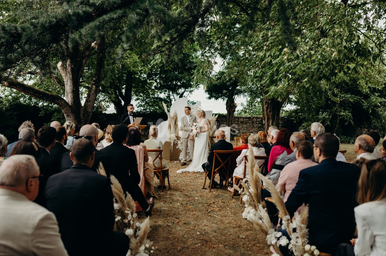 Cérémonie de mariage en plein air, avec des invités assis et un couple échangeant leurs vœux devant un officiant.