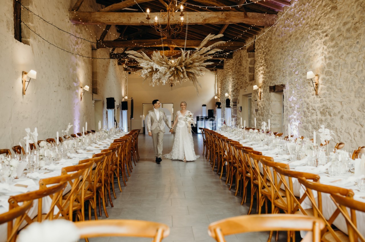 Un couple en tenue de mariage marche au centre d'une salle décorée avec des tables alignées et des guirlandes lumineuses.
