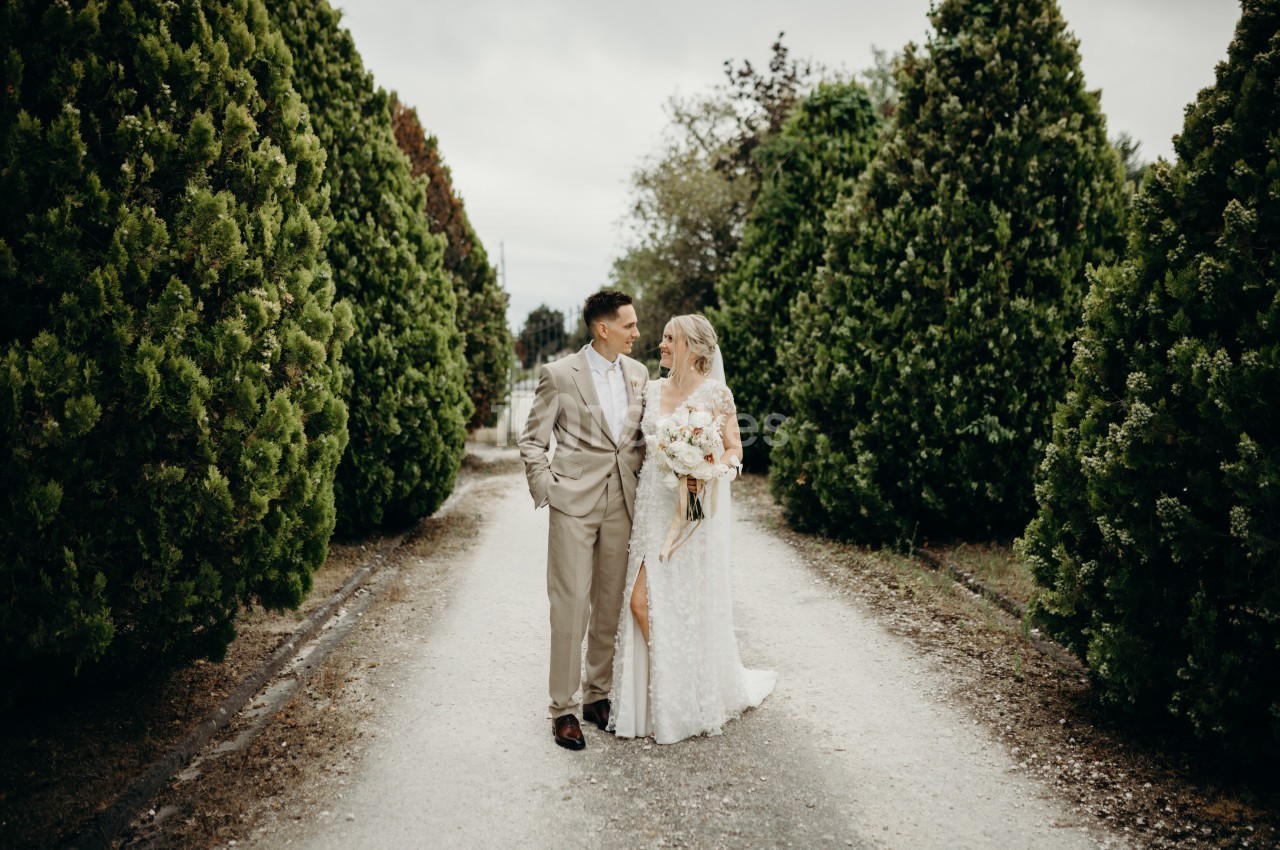 Un couple en tenue de mariage marche sur une allée bordée de grands arbres verts par temps nuageux.