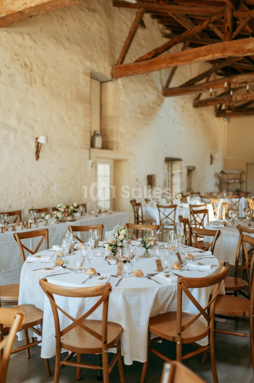 Salle de réception lumineuse avec tables rondes dressées, nappes blanches, chaises en bois et décorations florales.
