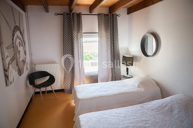 Chambre lumineuse avec deux lits simples, rideaux à motifs, tableau mural et miroir rond au mur.