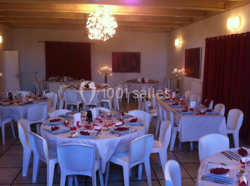 Salle décorée pour un événement, avec des tables rondes dressées, nappes blanches et accents rouges, éclairage chaleureux.