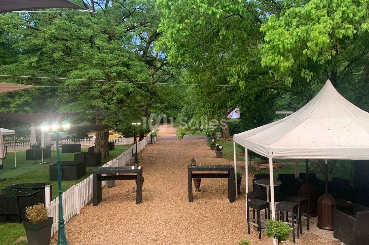 Allée en gravier bordée de tables, chaises et parasols, entourée de verdure et clôtures blanches.