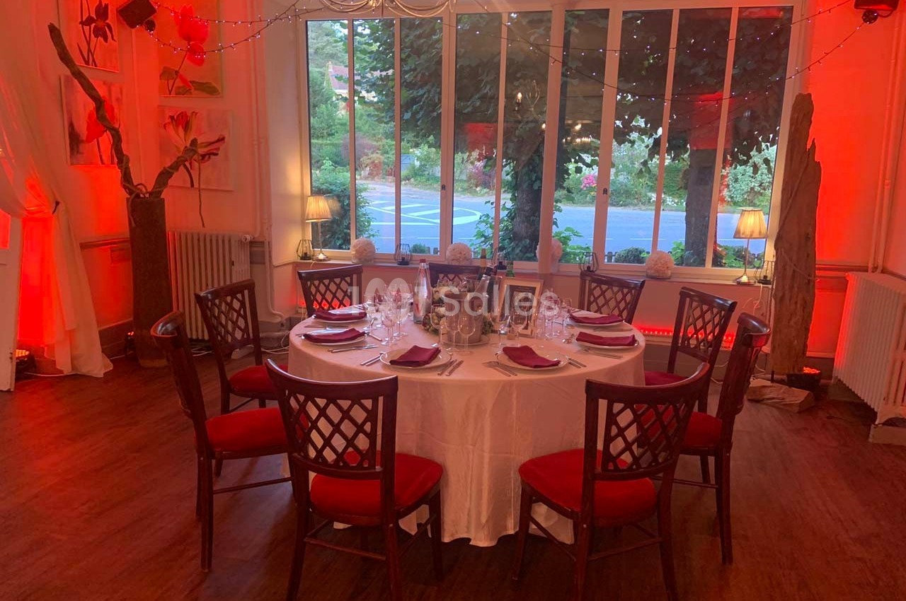 Salle décorée avec une table ronde dressée pour un repas, entourée de chaises en bois avec coussins rouges.