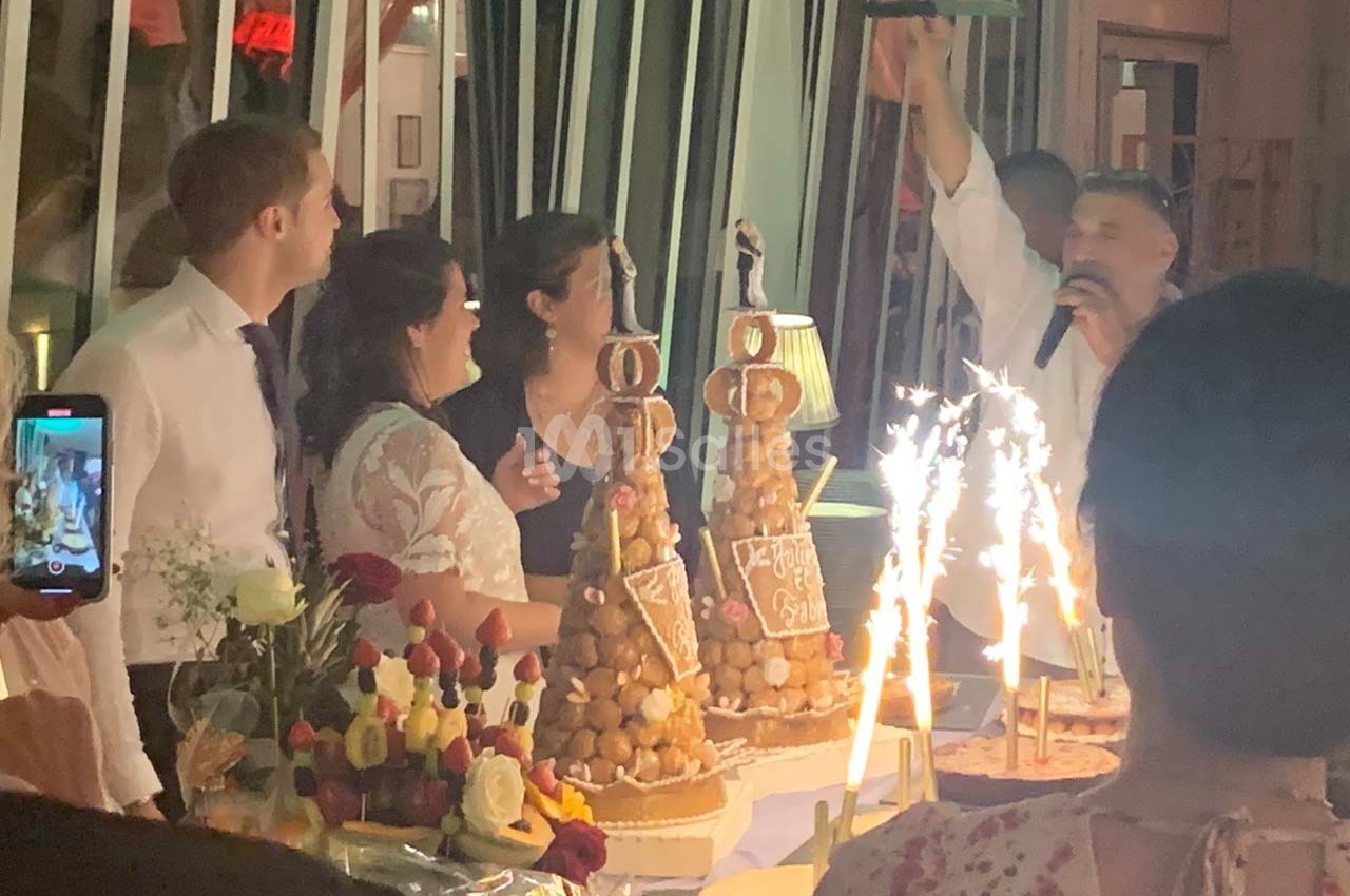Un groupe de personnes autour de pièces montées et desserts, avec des étincelles et un animateur parlant au micro.