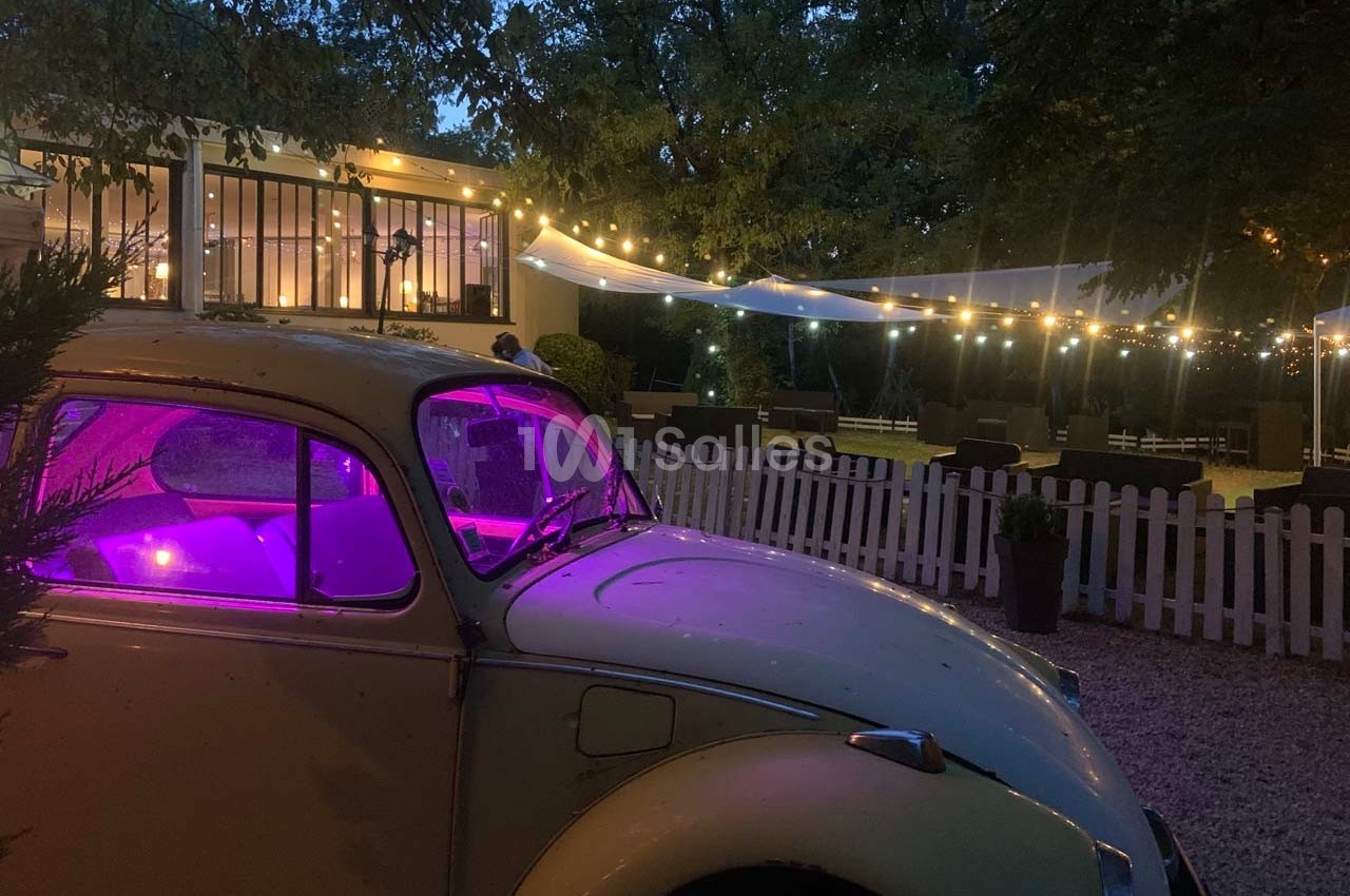 Voiture ancienne éclairée en violet devant une terrasse illuminée et décorée de guirlandes lumineuses, en soirée.