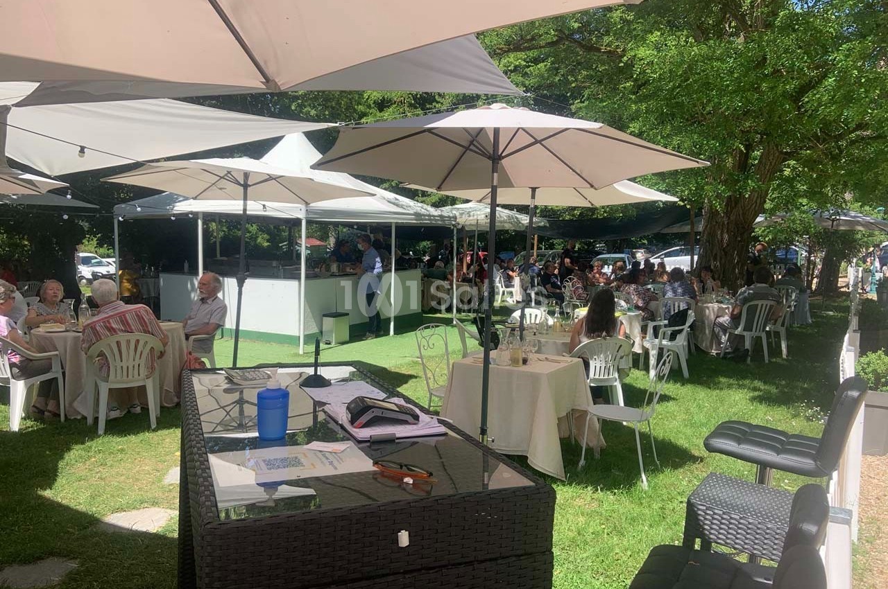 Des personnes déjeunent en plein air sous des parasols dans un espace verdoyant avec des tables et un bar en arrière-plan.