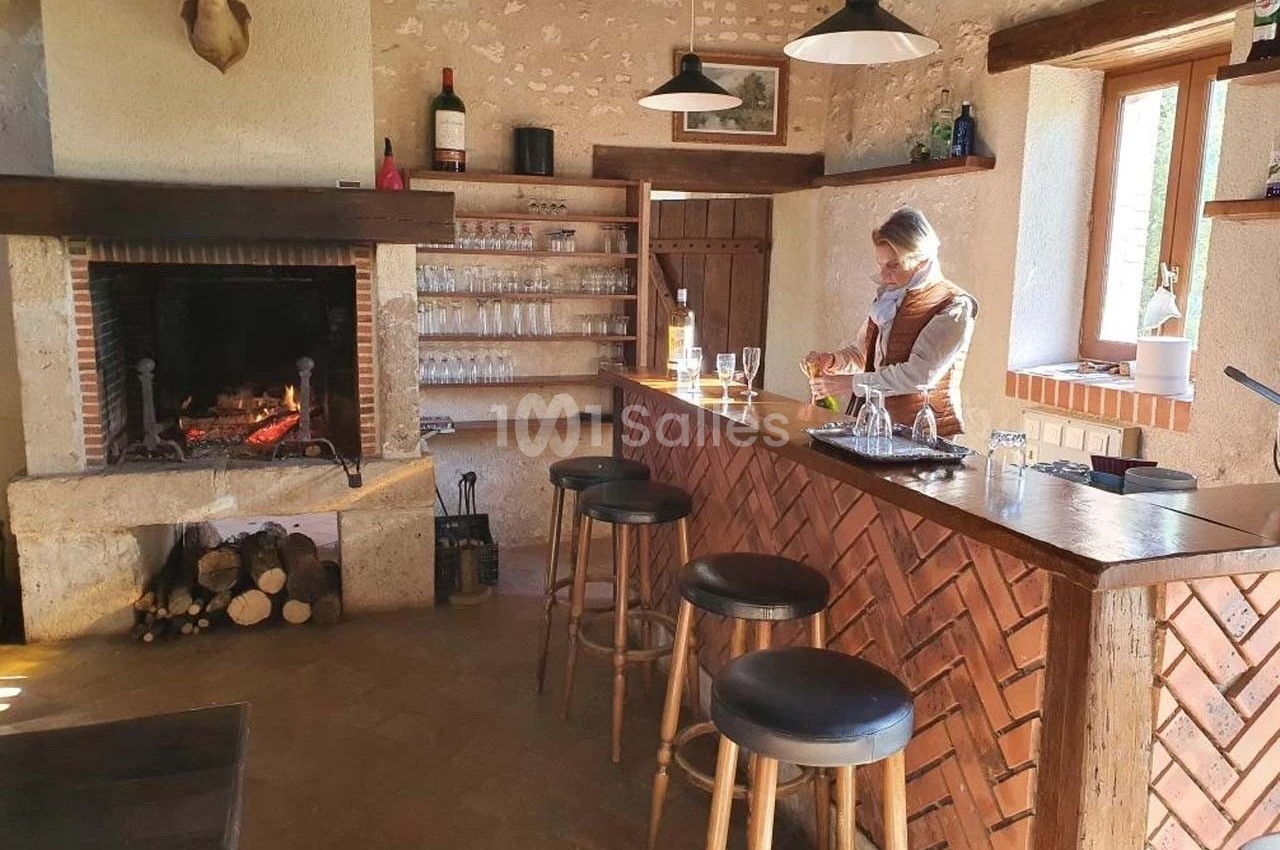 Une femme prépare des verres derrière un comptoir en bois dans une pièce rustique avec cheminée et étagères.