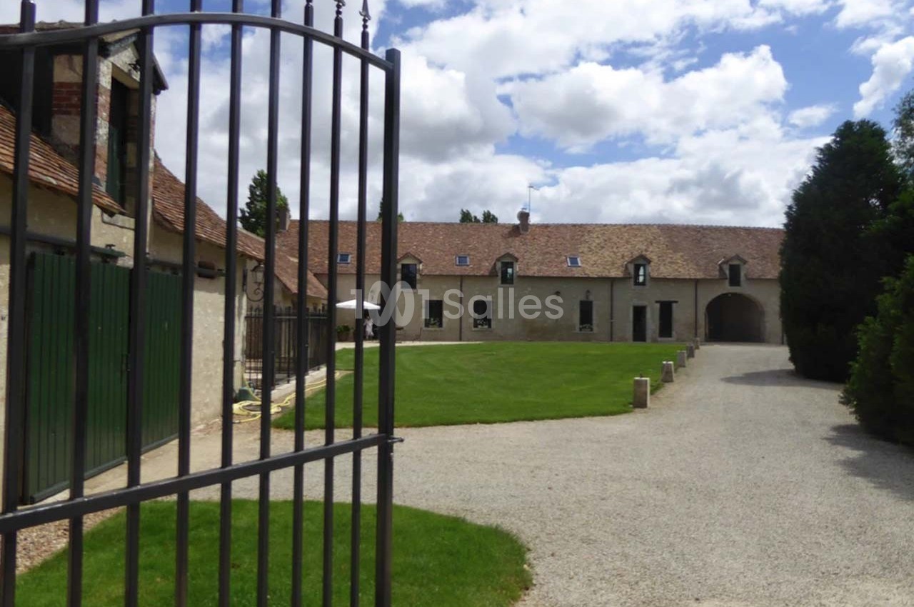 Cour d'une grande bâtisse en pierre avec toit en tuiles, vue depuis un portail en fer forgé, ciel partiellement nuageux.