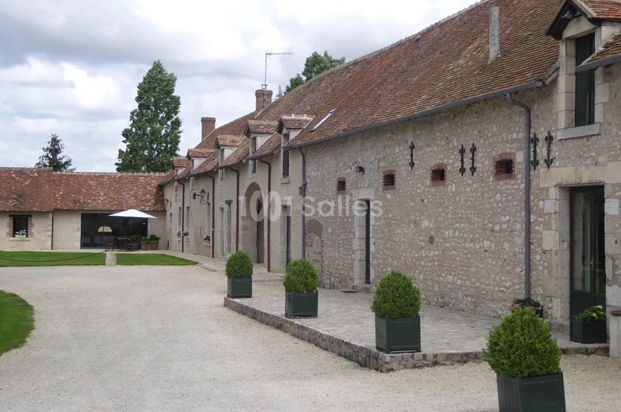 Bâtiment en pierre avec toit en tuiles, fenêtres mansardées et allée gravillonnée bordée de bacs à plantes.