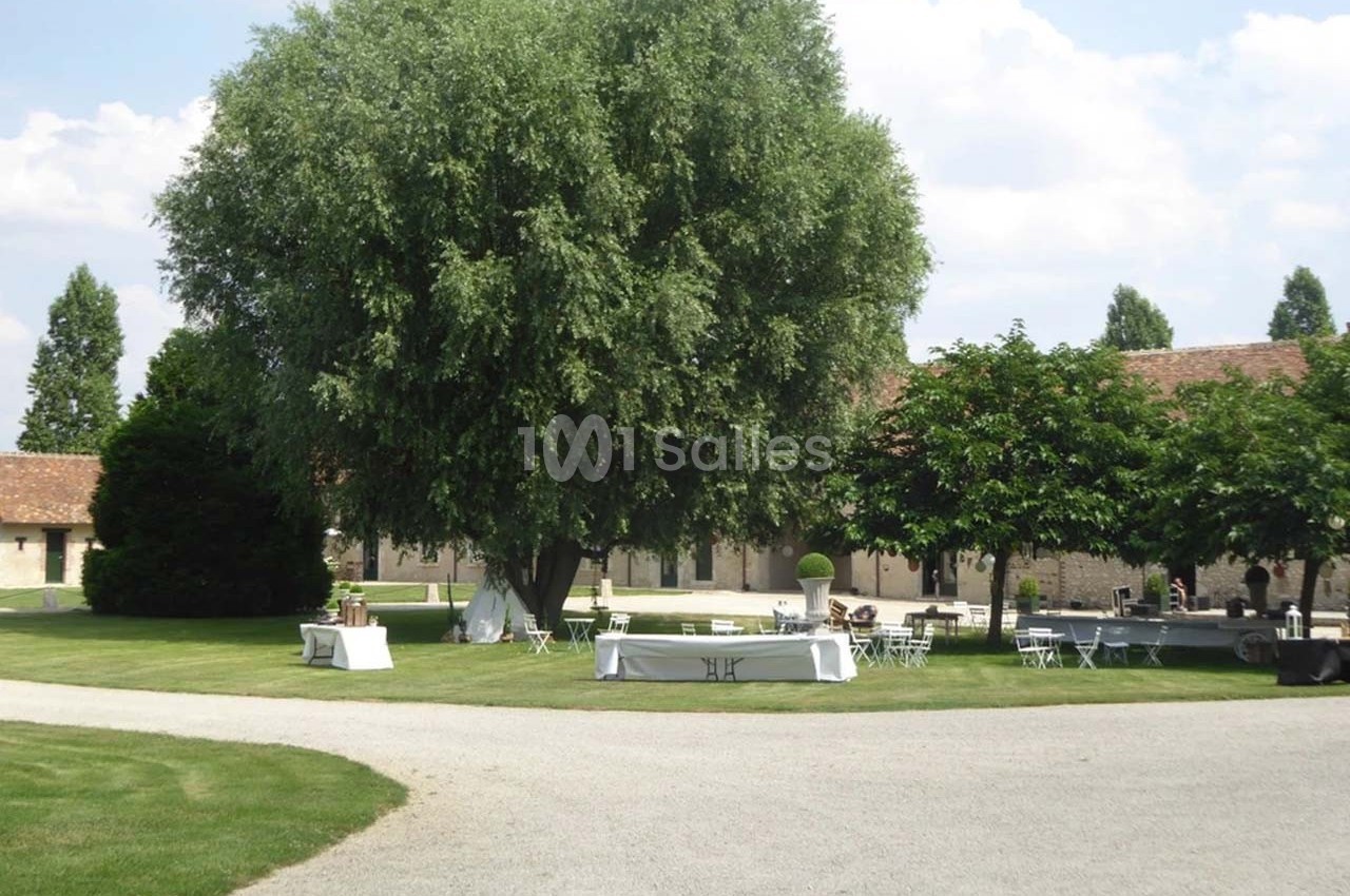 Espace extérieur avec pelouse, grands arbres, tables et chaises blanches disposées devant un bâtiment en pierre.