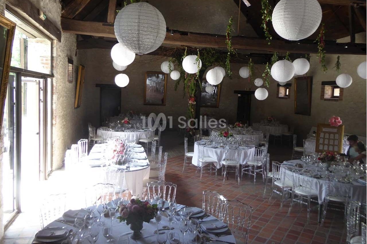 Salle de réception décorée avec des lanternes blanches suspendues, tables dressées et chaises transparentes.