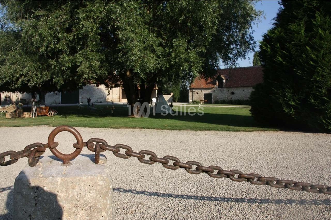 Chaîne en métal attachée à un poteau en pierre, avec en arrière-plan un jardin, des arbres et des bâtiments anciens.