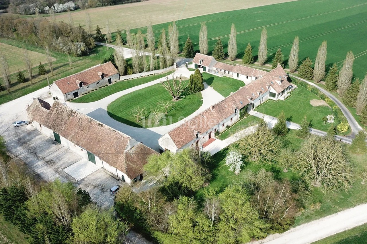 Vue aérienne d'une grande ferme en U avec bâtiments en pierre, toits en tuiles et cour centrale entourée de verdure.