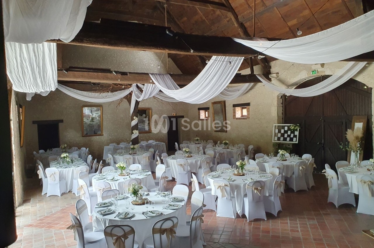 Salle de réception avec tables rondes décorées, nappes blanches et plafonds ornés de voilages suspendus.