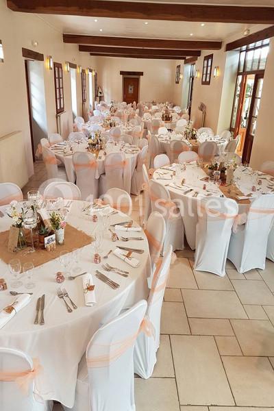 Salle de réception décorée pour un événement, avec des tables rondes dressées et des chaises blanches ornées de rubans.