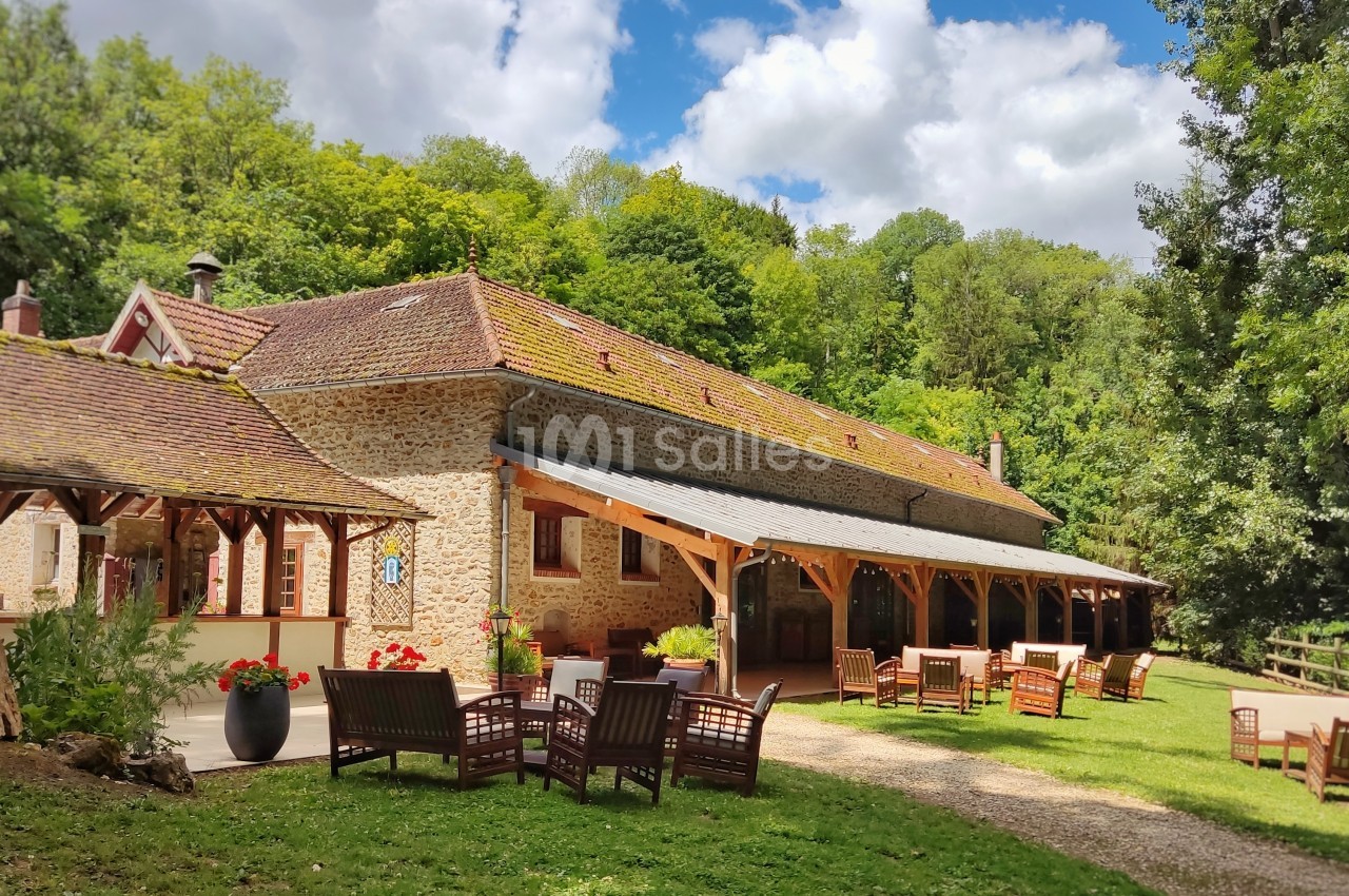 Bâtiment en pierre avec toit en tuiles, terrasse aménagée et mobilier de jardin, entouré de verdure sous un ciel…