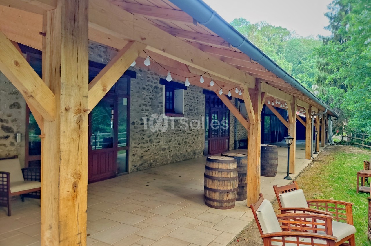 Terrasse couverte en bois avec des tonneaux décoratifs, des fauteuils et un mur en pierre, entourée de verdure.