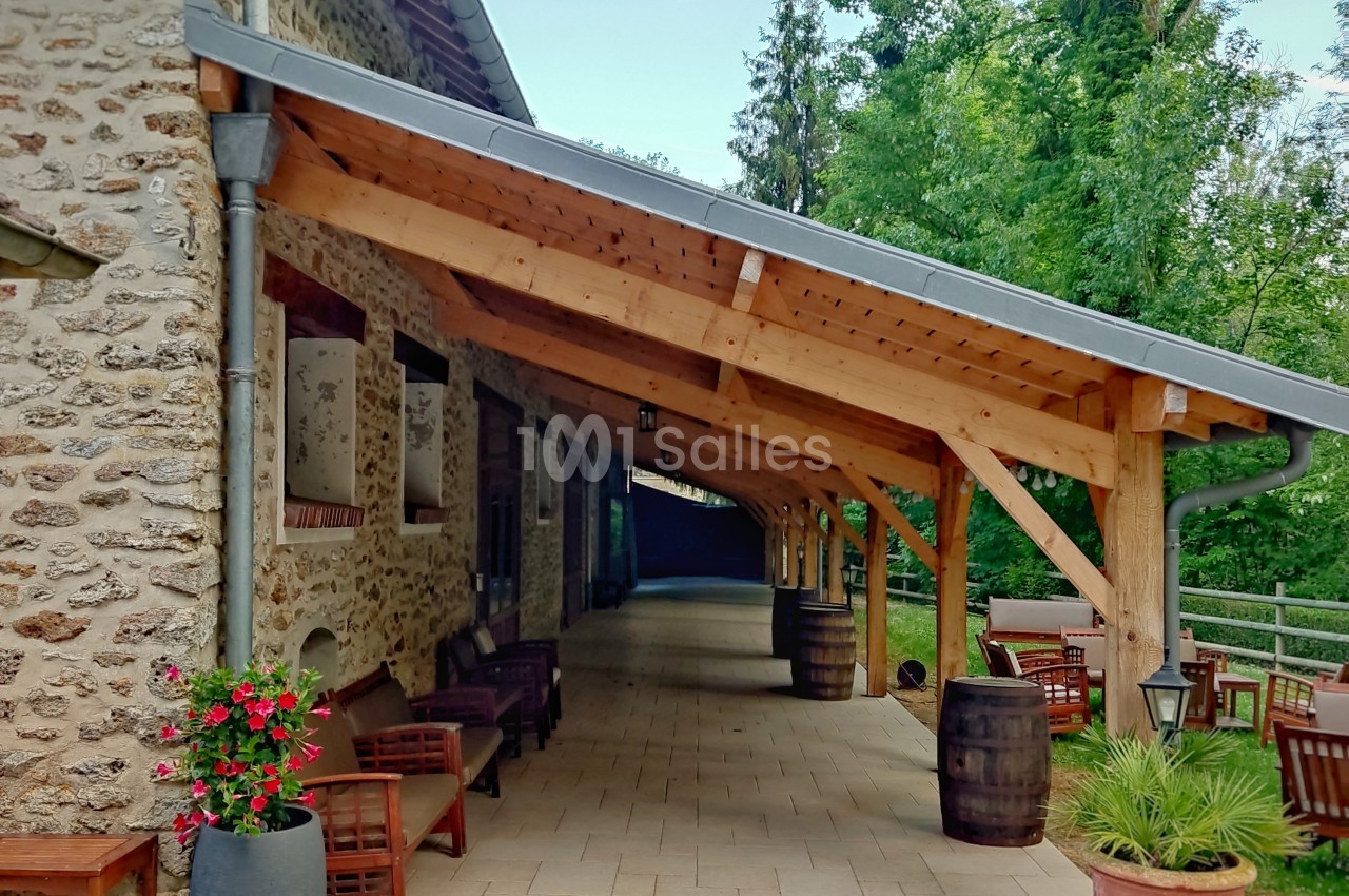 Terrasse couverte en bois attenante à un bâtiment en pierre, avec des tables, des chaises et des plantes en pot.