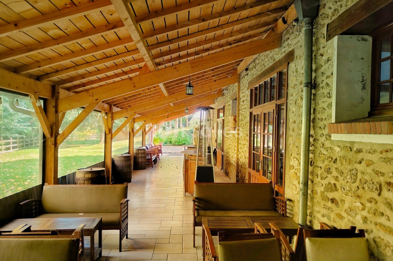 Terrasse couverte en bois avec mobilier de jardin, jouxtant un bâtiment en pierre et donnant sur un espace vert.