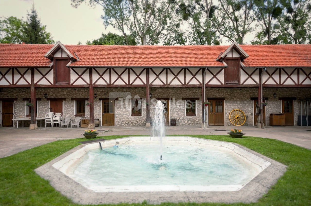 Cour intérieure avec une fontaine centrale, entourée de bâtiments en pierre et bois à toit rouge, bordée de verdure.