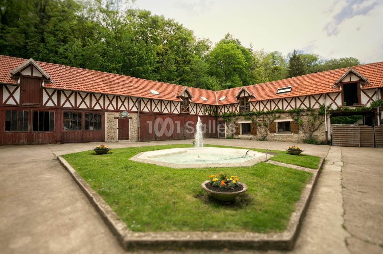 Cour intérieure pavée avec une fontaine centrale, entourée de bâtiments à colombages et toits en tuiles rouges.