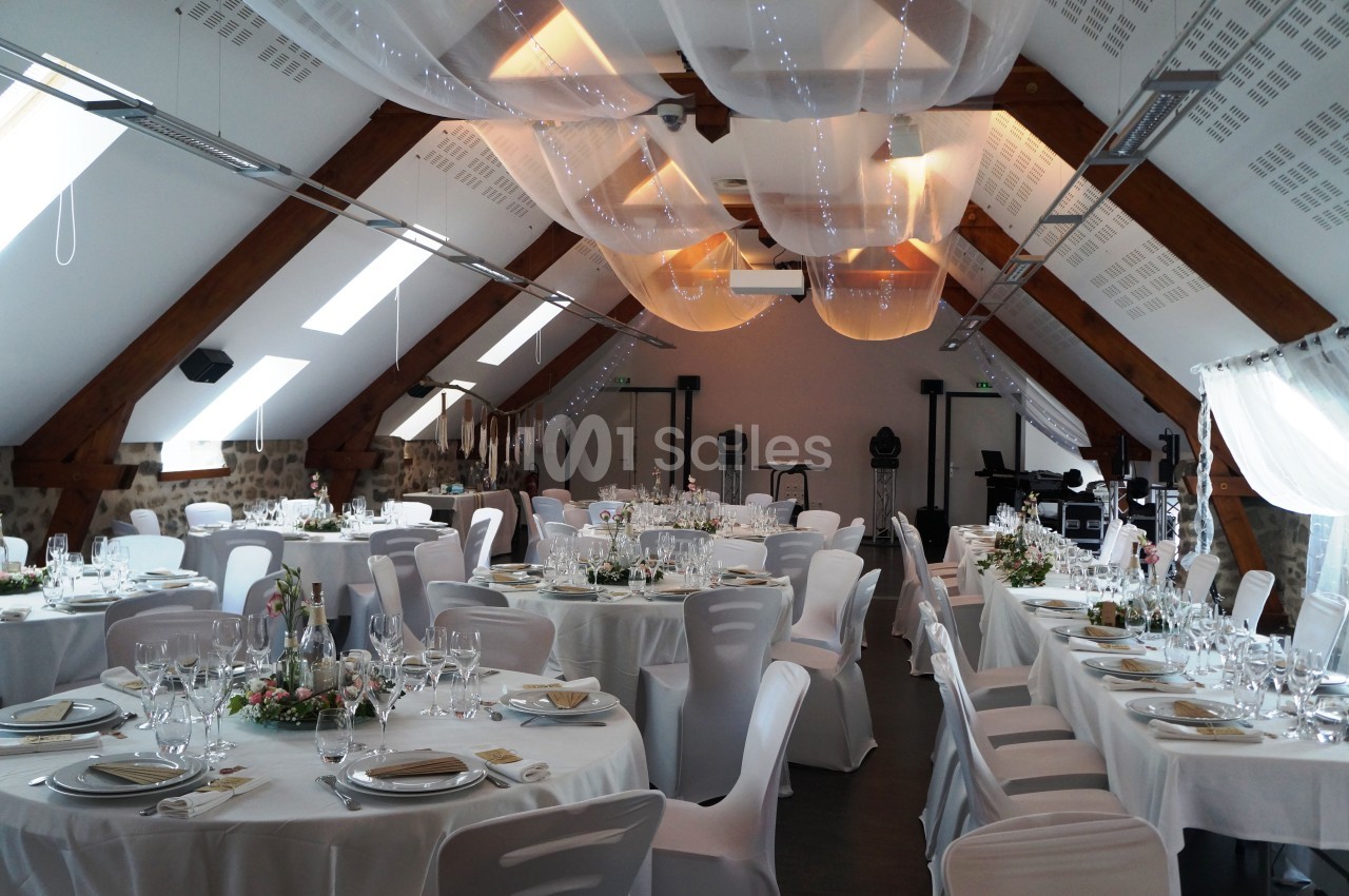 Salle de réception décorée avec des tables rondes dressées, des chaises blanches et un plafond orné de voilages lumineux.