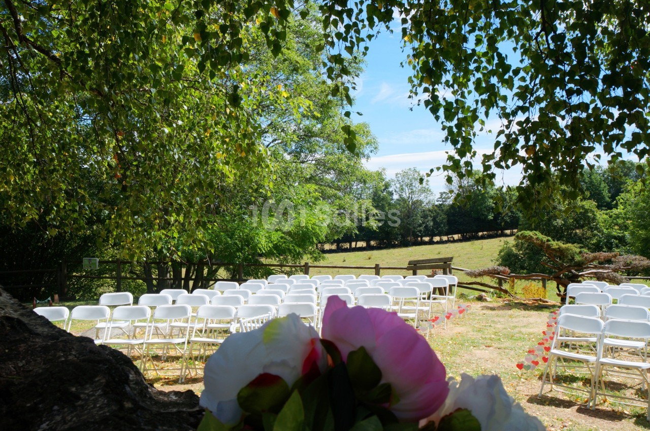 Chaises blanches alignées en extérieur sous des arbres, avec un bouquet de fleurs au premier plan et un champ en arrière…