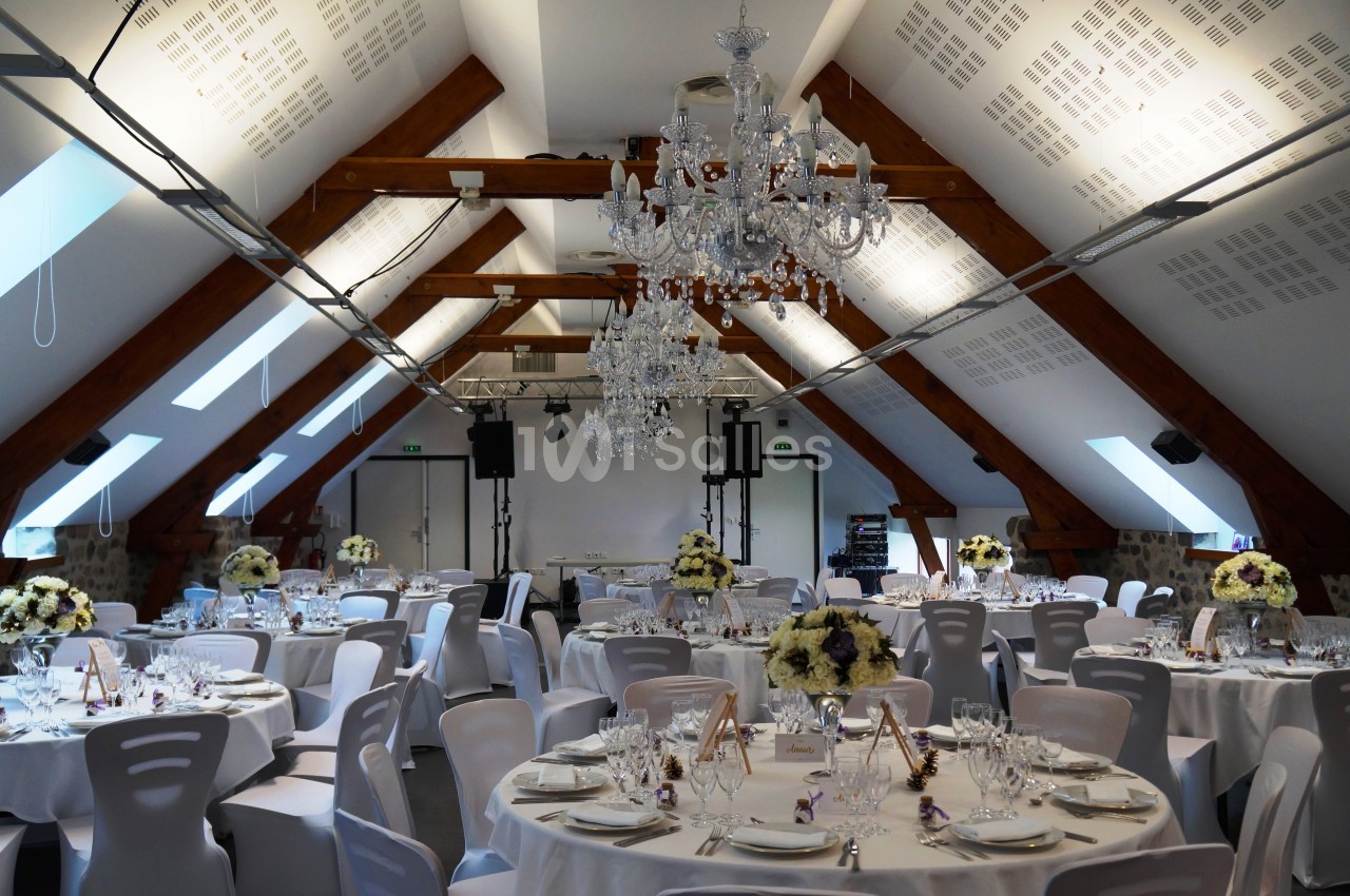 Salle de réception décorée avec des tables rondes dressées, des chaises blanches et des lustres suspendus.