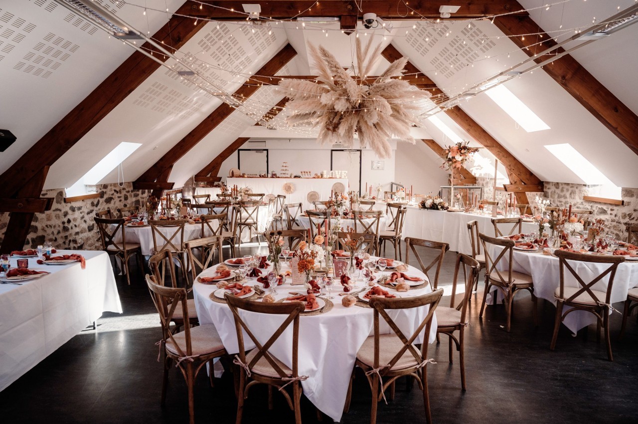 Salle de réception décorée pour un mariage, avec tables rondes, nappes blanches et ornements floraux suspendus.