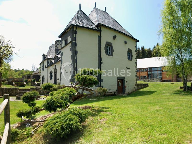 Bâtiment ancien en pierre avec toit en ardoise, entouré d'un jardin verdoyant et d'arbres, sous un ciel dégagé.