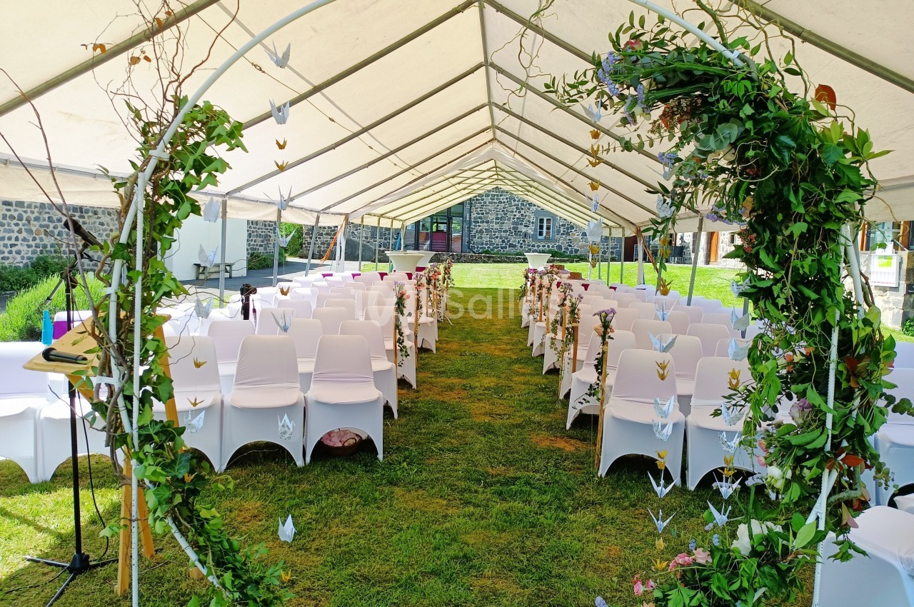 Allée centrale décorée pour une cérémonie sous une tente, entourée de chaises blanches et d'ornements floraux.