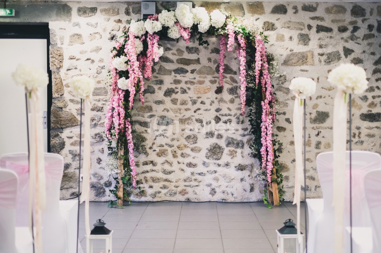 Arche florale décorée de fleurs blanches et roses devant un mur en pierre, dans une salle de cérémonie.