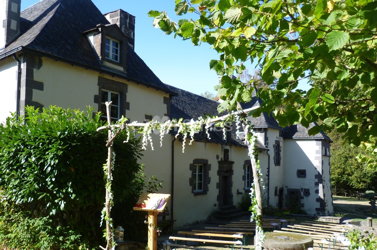 Façade d'une maison en pierre avec toit en ardoise, entourée de verdure et ornée d'une arche fleurie.