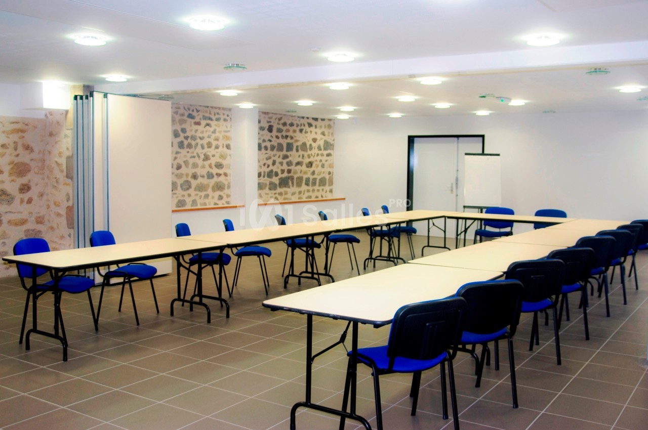 Salle de réunion lumineuse avec tables disposées en U, chaises bleues et murs en pierre apparente.