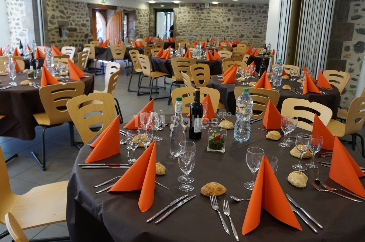 Salle de réception avec tables rondes dressées, nappes marron, serviettes orange et couverts disposés.