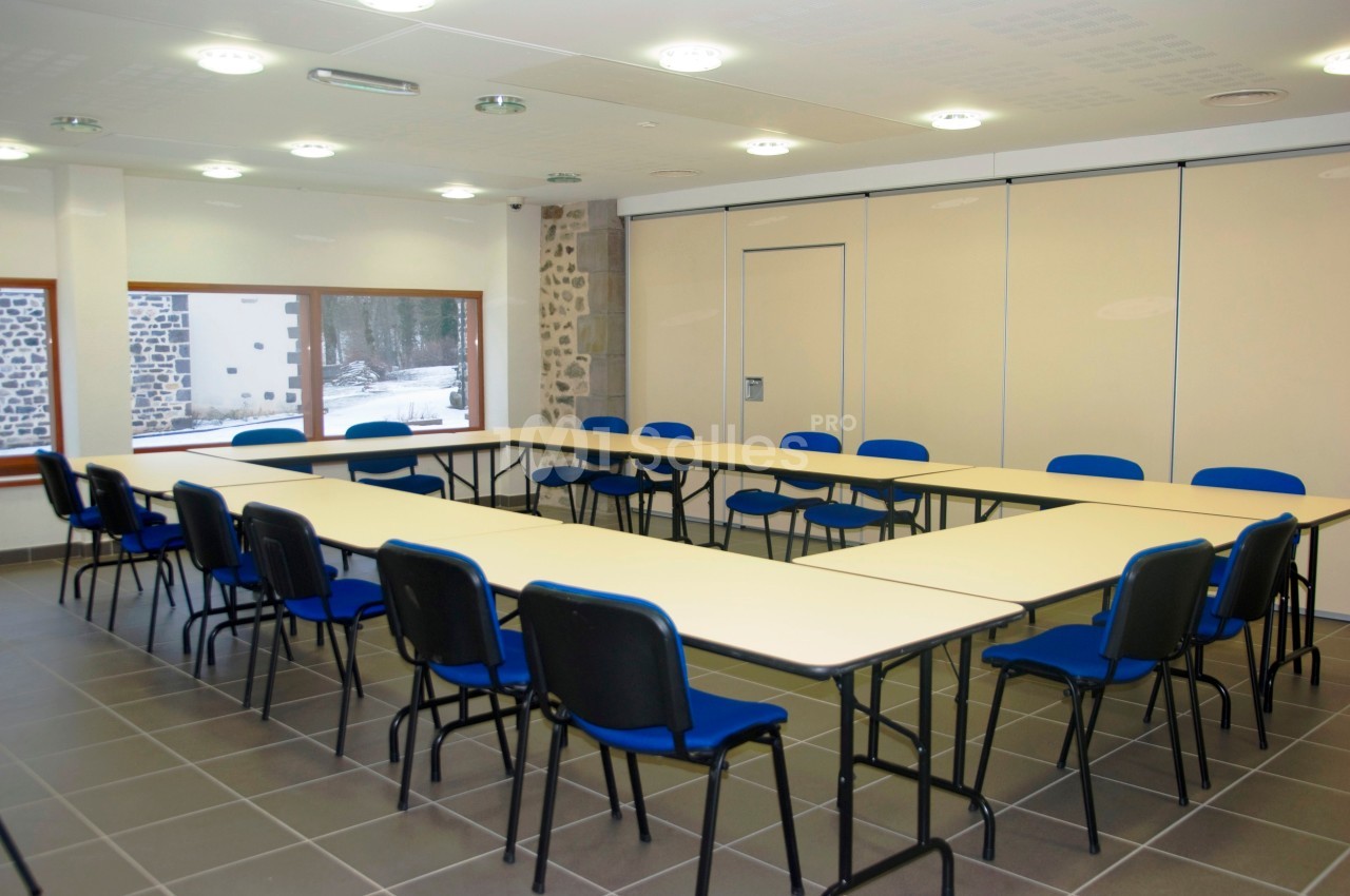 Salle de réunion lumineuse avec des tables disposées en U et des chaises bleues sur un sol carrelé.