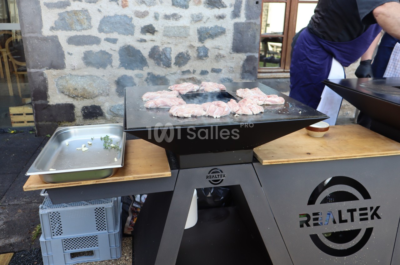 Cuisson de morceaux de viande sur une plancha en extérieur, avec un mur en pierre en arrière-plan.