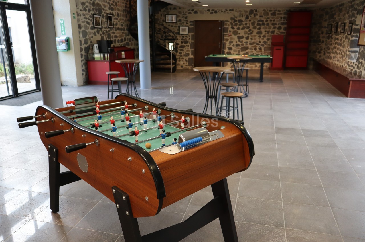 Baby-foot en bois dans une salle spacieuse avec tables, chaises et murs en pierre apparente.