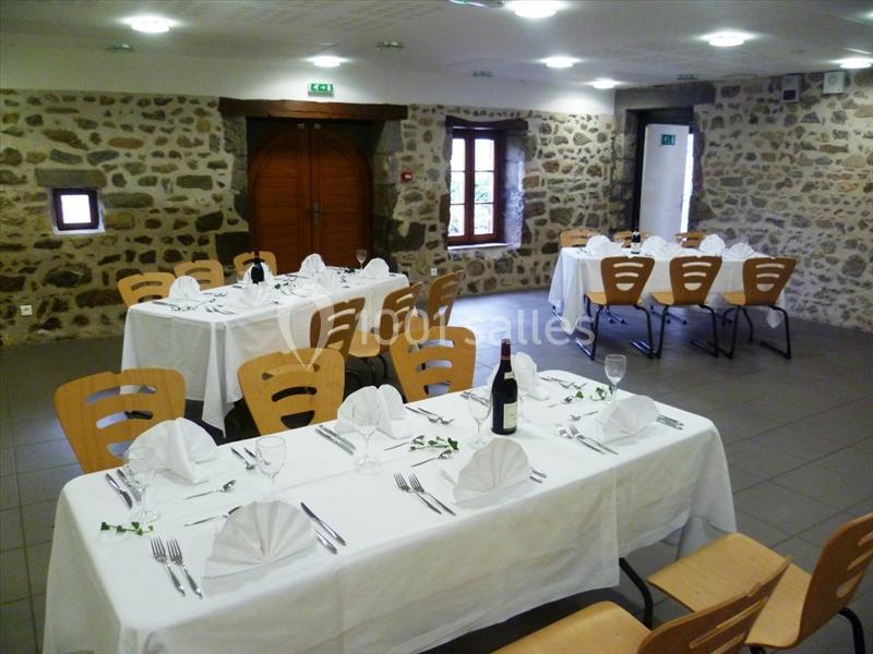 Salle en pierre aménagée avec des tables dressées pour un repas, nappes blanches et chaises en bois.
